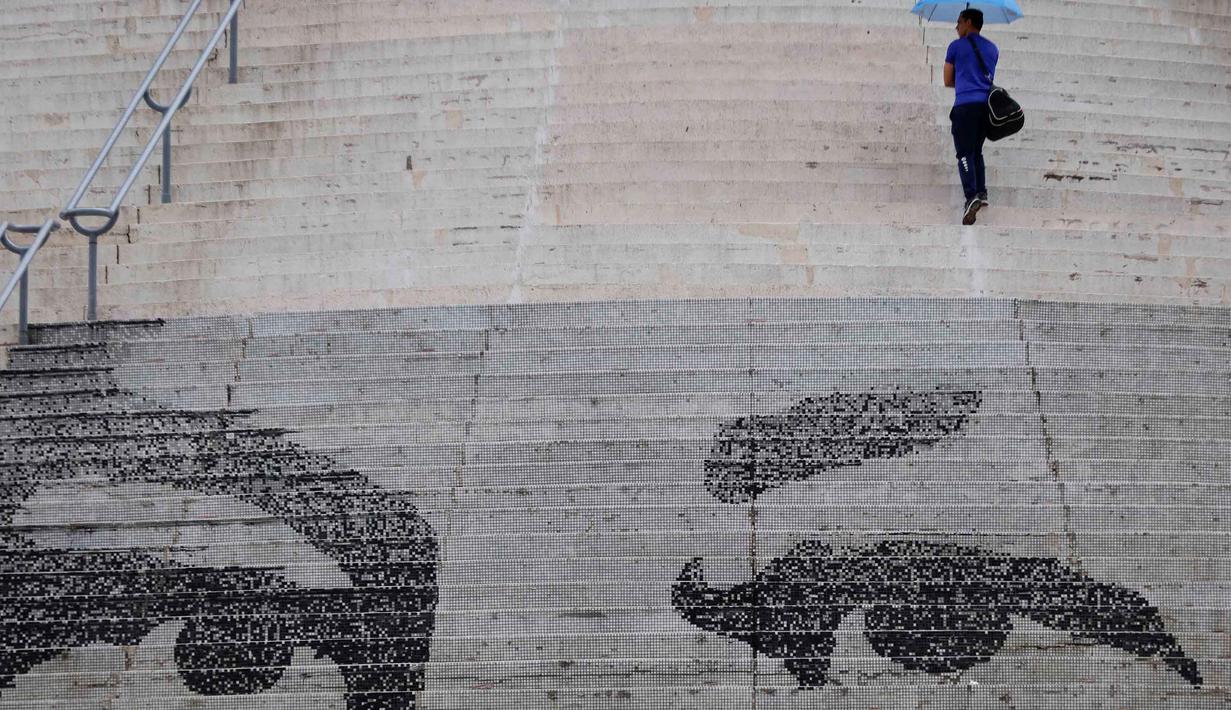 Pemerintah Amerika Serikat (AS) mendakwa Nicolas Maduro terlibat dalam konspirasi narkoterorisme internasional, penyelundupan kokain, dan pelanggaran senjata api. Tampak dalam foto, seorang pria berjalan sambil membawa payung melewati mosaik yang menggambarkan mata mendiang Presiden Venezuela Hugo Chavez di tangga Lapangan Calvario selama pawai dukungan untuk mantan Presiden Venezuela Nicolas Maduro, yang diselenggarakan oleh para pekerja di Caracas pada 14 Januari 2026. (Pedro MATTEY/AFP)