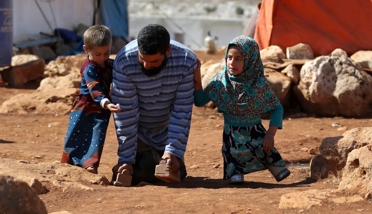 Maya Mohammad Ali Merhi (kanan) berjalan dengan sang ayah (tengah) dan seorang anak lain di sebuah kamp orang terlantar di Provinsi Idlib, Suriah, Rabu (20/6). Maya dan ayahnya lahir tanpa tubuh bagian bawah. (Aaref Watad/AFP)