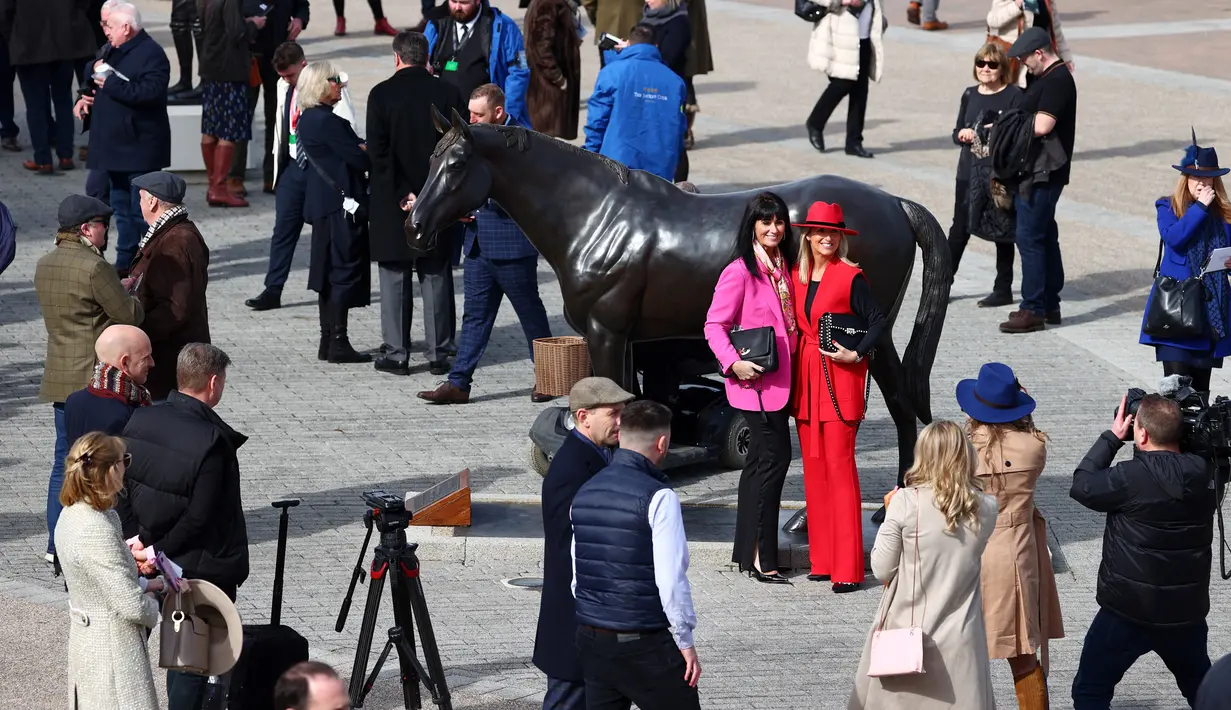 Dua wanita berpose di samping patung "Best Mate" saat orang-orang tiba pada hari pertama Festival Cheltenham di Cheltenham Racecourse, di Cheltenham, Inggris (15/3/2022). Cheltenham adalah pertemuan berbasis pacuan kuda dalam kalender balap Perburuan Nasional di Inggris Raya. (AFP/Adrian Dennis)