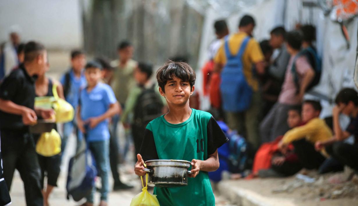 Seorang anak laki-laki Palestina yang mengungsi membawa sepanci makanan yang dikumpulkan dari dapur amal ke tempat penampungan keluarganya, di kamp pengungsi Bureij, Jalur Gaza Tengah pada Senin 18 Mei 2026. (Eyad Baba/AFP)