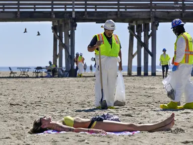 Pekerja membersihkan minyak dari pasir dekat seorang wanita yang berbaring di bawah sinar matahari di Pantai Newport, California, Amerika Serikat, Selasa (5/10/2021). Kebocoran pipa menyebabkan tumpahan minyak di lepas pantai California. (Jeff Gritchen/The Orange County Register via AP)