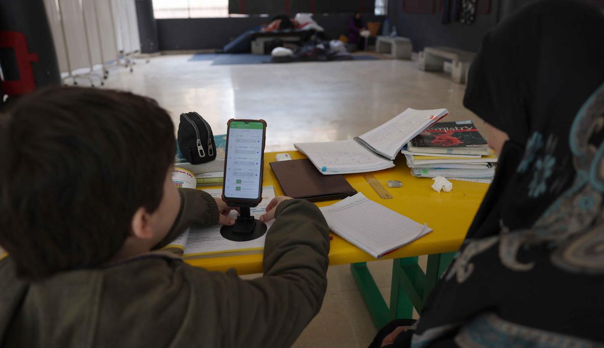 Banyak gedung sekolah di Lebanon dialihfungsikan menjadi pusat penampungan bagi warga yang mengungsi. Tampak dalam foto, seorang wanita Lebanon yang mengungsi duduk di samping seorang anak, mengawasi pelajaran daringnya melalui telepon seluler di sebuah sekolah negeri yang diubah menjadi tempat penampungan di kota Dekwaneh, utara Beirut, pada Rabu 25 Maret 2026. (Anwar AMRO/AFP)