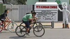 Pesepeda melintas di depan seni instalasi yang menggambarkan tenaga medis memegang peti jenazah korban COVID-19 di Kemang 1, Jakarta Selatan, Minggu (16/8/2020). Seni instalasi itu dipajang guna memberi peringatan kepada warga untuk tetap waspada terhadap COVID-19. (Liputan6.com/Herman Zakharia)