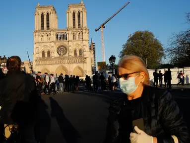 Seorang perempuan melintasi orang-orang yang berhenti di depan Katedral Notre Dame saat kembali membunyikan lonceng utamanya di Paris, Rabu (15/4/2020). Lonceng utama katedral kembali dibunyikan setelah satu tahun mengalami kebakaran yang sampai mengejutkan seluruh warga dunia. (AP/Christophe Ena)