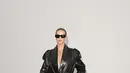 Rosie Huntington-Whiteley memilih trench coat kulit hitam mengilap bermodel oversized, dipadukan kacamata hitam besar, kalung rantai, dan heels bertali hitam. [dok. Burberry]