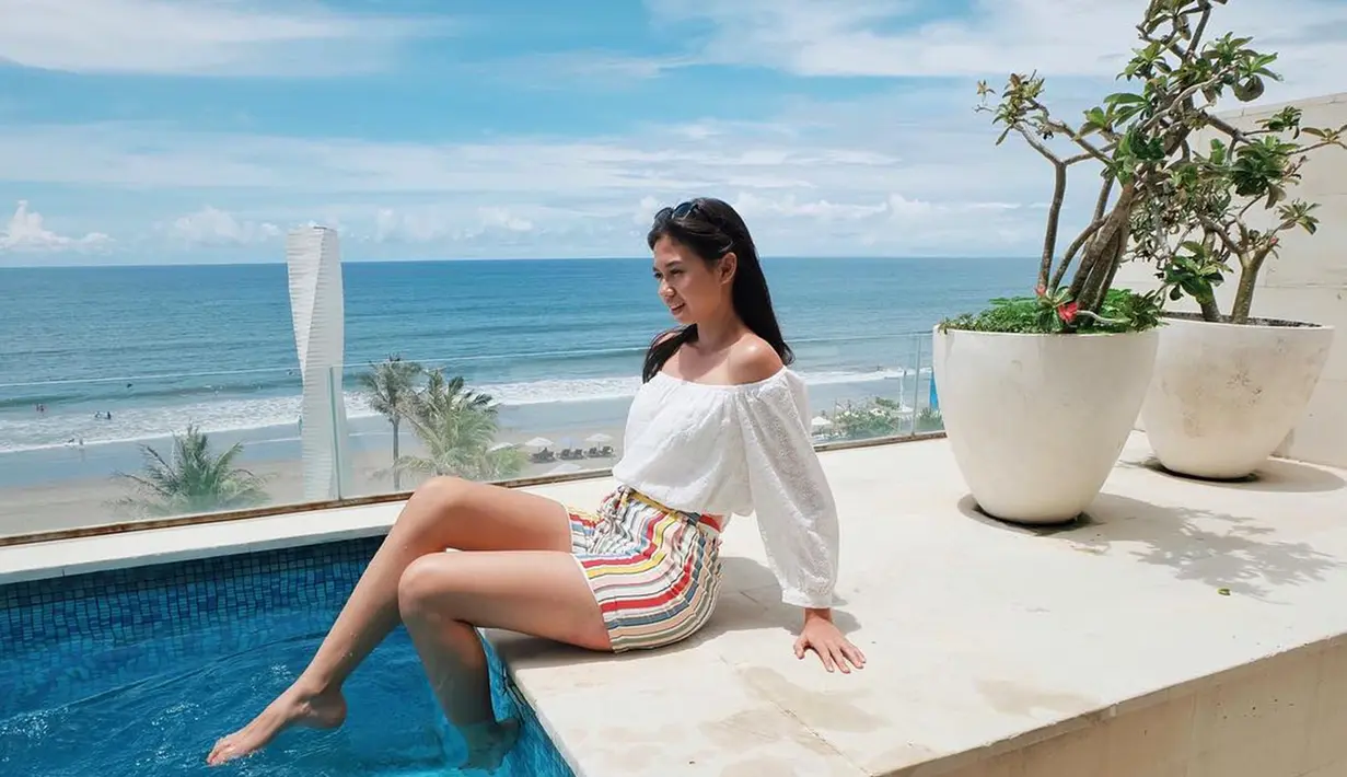 Yuki Kato tampak bahagia saat bermain air di sebuah kolam renang hotel di Bali. Menggunakan pakaian putih dan celana garis-garis warna-warni, latar belakang lautan dan langit biru menambah keindahan pemandangan.(Liputan6.com/IG/@yukikt)