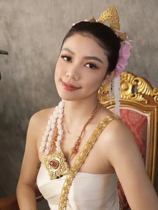 Pakaian adat Chut Thai yang dikenakan Regina terdiri dari tube top putih yang dipadukan dengan kain tenun coklat keemasan. Dihiasi dengan sabuk emas tradisional yang berkilau, penampilannya semakin memikat hati para penggemar. Aksesori emas yang melengkapi penampilannya, seperti mahkota kecil, anting, gelang, dan kalung dengan liontin besar, semakin memperkuat kesan kerajaan dan elegan.  (Liputan6.com/IG/@reginaaphx)