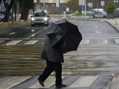 Seorang pria memegang payungnya melawan hujan yang disertai angin saat menyeberang jalan di Alges, tepat di luar Lisbon, Selasa (13/12/2022). Badai Atlantik menghantam semenanjung Iberia, meninggalkan jejak kehancuran pada Selasa, terutama di Ibu kota Portugal,  Lisbon, sebelum pindah ke timur ke Spanyol.  (AP Photo/Armando Franca)