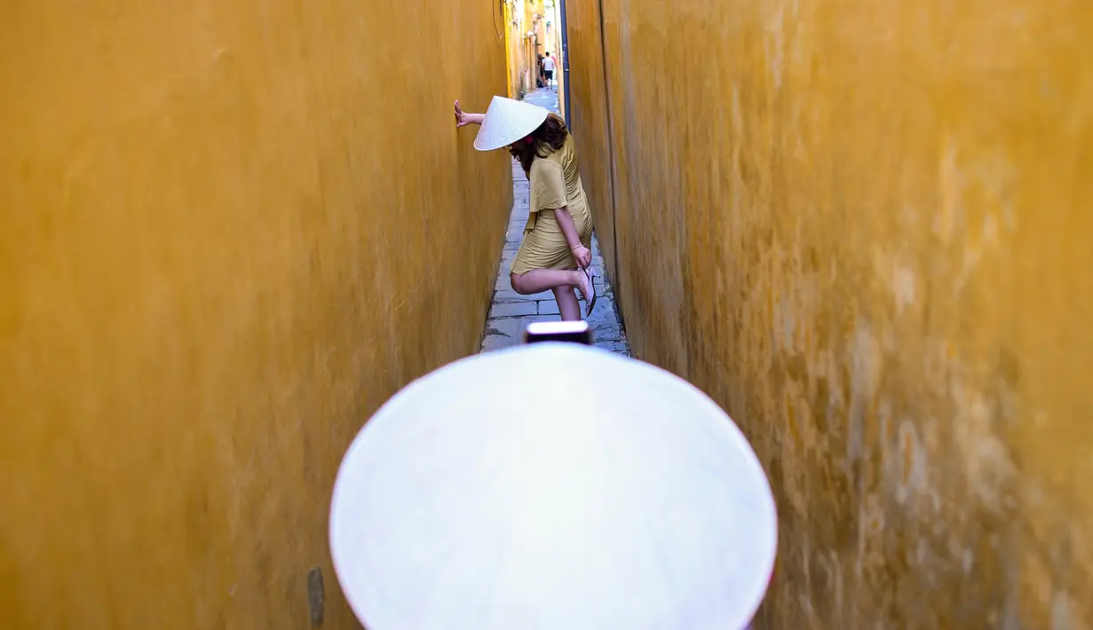 Seorang wanita mengenakan topi tradisional 'Non La' sambil berpose di lorong yang dicat warna kuning di kota tua Hoi An, 14 September 2019. Kota Tua Hoi An merupakan destinasi wisata populer di Vietnam yang mendapat gelar sebagai salah satu situs warisan dunia dari UNESCO. (Manan VATSYAYANA/AFP)