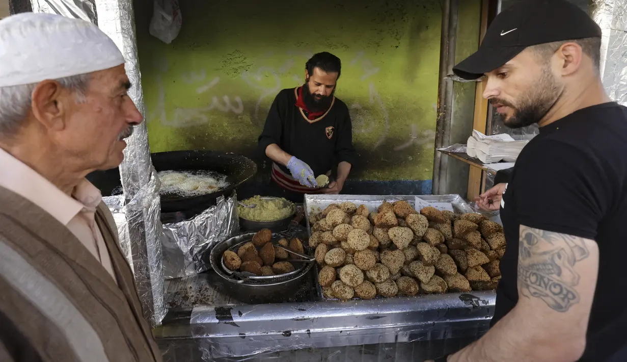 Pelanggan menunggu saat pedagang membuat falafel untuk hidangan berbuka puasa, selama bulan suci Ramadhan di pusat kota Hebron di Tepi Barat yang diduduki (9/4/2022). Makanan ini dibuat dari kacang Arab yang digiling dan kemudian dipadatkan dan dibuat bola-bola kecil lalu digoreng. (AFP/Hazem Bader)