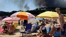 Wisatawan berjemur sambil menyaksikan asap hitam yang membumbung akibat kebakaran hutan dari pinggir pantai Lavandou, Rabu (26/7). Prancis juga telah meminta bantuan negara tetangga di Uni Eropa untuk membantu memadamkan kebakaran itu. (AP/Claude Paris)
