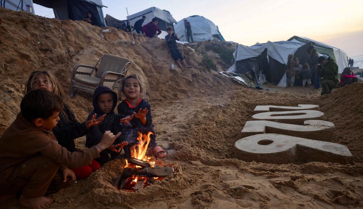 Dan, lebih dari 171.222 orang mengalami luka-luka, banyak di antaranya menderita cedera permanen yang mengubah hidup. Tampak dalam foto, anak-anak menghangatkan diri di dekat api unggun di samping patung pasir karya seniman Yazed Abo Jarad yang menggambarkan tahun mendatang di Deir El-Balah, Jalur Gaza tengah pada 30 Desember 2025. (Bashar Taleb/AFP)
