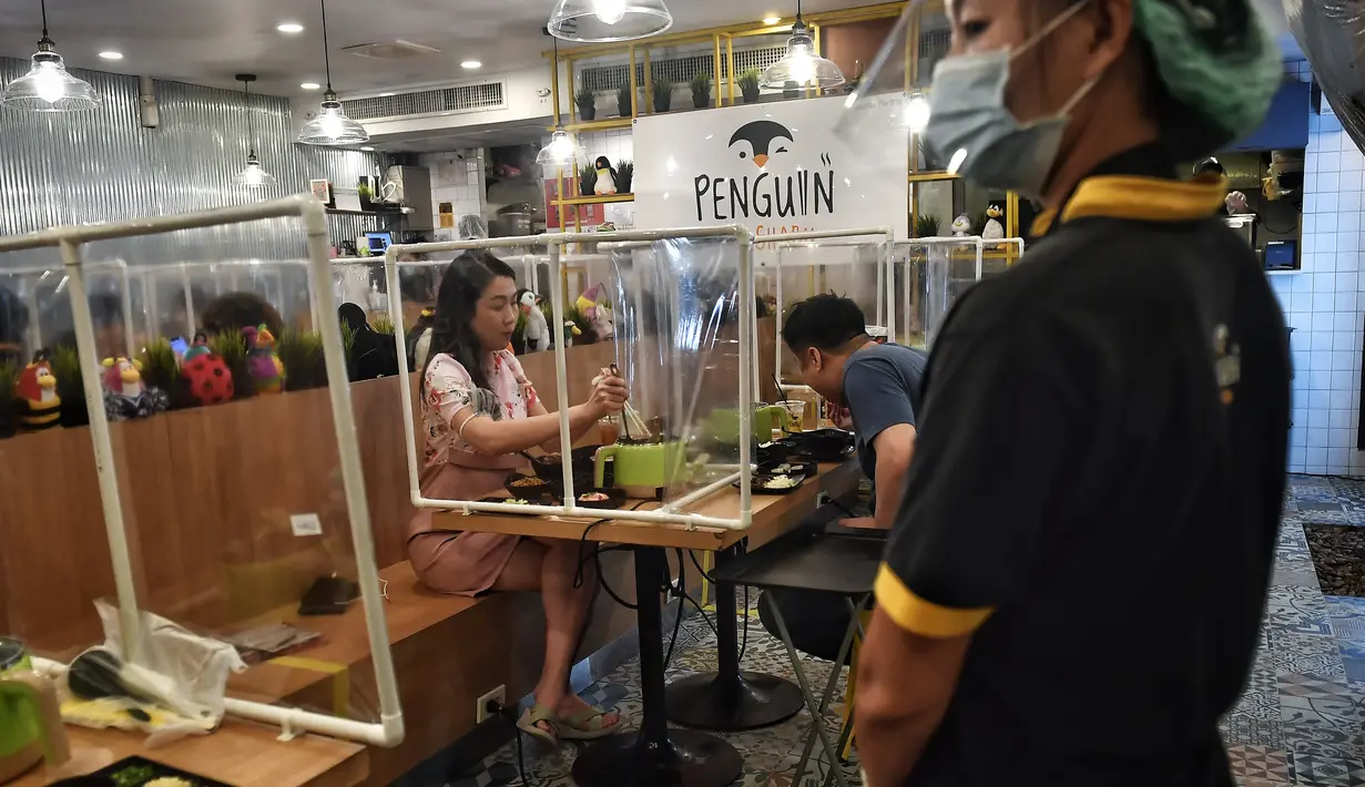 Pramusaji dengan pelindung wajah terlihat saat pelanggan menikmati makanan di restoran hotpot Penguin Eat Shabu di Bangkok, 5 Mei 2020. Restoran yang memasang pembatas plastik itu kembali dibuka setelah Thailand memperlonggar lockdown dengan menerapkan social distancing. (Lillian SUWANRUMPHA/AFP)