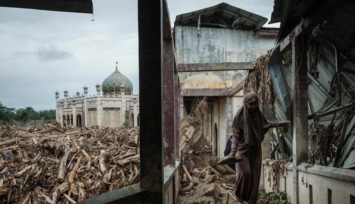Seorang siswi berjalan di atas bangku untuk menghindari lumpur karena tumpukan pohon tumbang yang tersapu banjir bandang masih berada di Pesantren Darul Mukhlisin dan masjid yang terhubung dengannya di Aceh Tamiang, pada Minggu 14 Desember 2025. Pemandangan memprihatinkan masih terlihat di Pondok Pesantren Darul Mukhlisin, Aceh Tamiang, Minggu (14/12/2025). (Yasuyoshi Chiba/AFP)