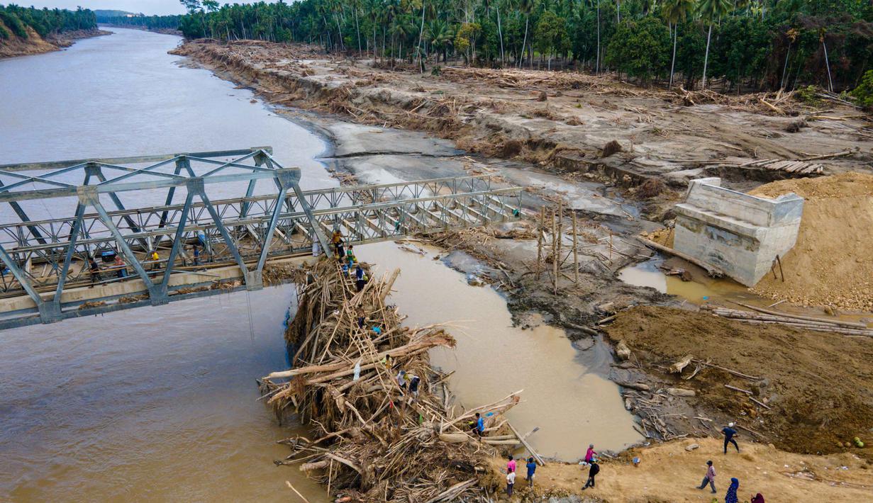 Struktur jembatan baja tersebut ditargetkan menjadi penghubung sementara agar mobilitas masyarakat dan distribusi logistik ke wilayah terdampak dapat kembali berjalan. Tampak foto udara menunjukkan warga memanjat puing-puing untuk menyeberangi sungai di jembatan yang baru dibangun yang menghubungkan Aceh dan Provinsi Sumatera Utara setelah hancur akibat banjir bandang di Sungai Peusangan di Distrik Bireuen, Provinsi Aceh, pada Selasa 9 Desember 2025. (CHAIDEER MAHYUDDIN/AFP)