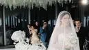 Ia tampil cantik dengan gaun putih saat memasuki altar. Gaun ini menggabungkan strapless dress dan lengan panjang transparan yang sopan   [Foto: Instagram/@sekisah.photo/@sekisah.sangjit].