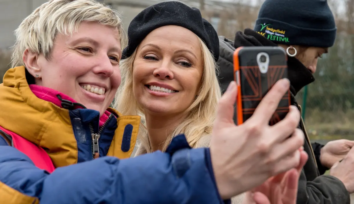 Pamela Anderson berselfie dengan relawan di pengungsian Grande-Synthe, Prancis (25/1). Pamela Anderson adalah seorang aktris yang terkenal berkat filmnya yang menceritakan penjaga pantai yang berjudul baywatch. (AFP/Philippe Huguen)