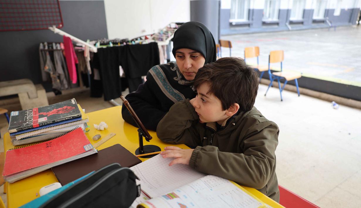 Seorang wanita Lebanon yang mengungsi duduk di samping seorang anak, mengawasi pelajaran daringnya melalui telepon seluler, di sebuah sekolah negeri yang diubah menjadi tempat penampungan di kota Dekwaneh, utara Beirut, pada Rabu 25 Maret 2026. Ketegangan di Lebanon semakin meningkat akibat eskalasi serangan udara yang dilakukan militer Israel. (Anwar AMRO/AFP)