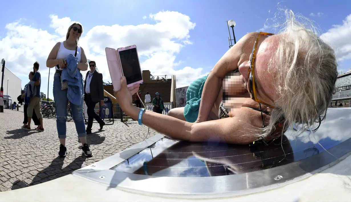 Lena Salmi mengisi baterai ponselnya saat berjemur di bangku panel surya di Helsinki, Finlandia, (9/7). Bangku panel surya dapat digunakan untuk mengisi ulang sepeda listrik dan perangkat mobile. (AFP Photo/Lehtikuva/Martti Kainulainen)