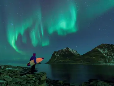 Seorang peselancar menatap Aurora sebelum beraksi di Lofoten Islands, Norwegia, (9/3/2018). Suhu udara minus 13°C dan suhu diatas air sekitar 4°C. (AFP/Oliver Morin)