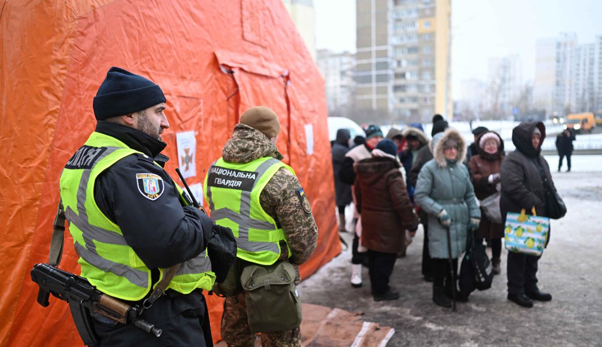 Mereka memasak dan mendistribusikan ribuan porsi makanan hangat kepada warga yang terdampak pemadaman listrik dan kesulitan mendapatkan akses makanan. Tampak dalam foto, petugas penegak hukum Ukraina berjaga-jaga saat warga setempat berkumpul untuk menerima makanan hangat yang dibagikan oleh sukarelawan dari badan amal makanan yang berbasis di Amerika Serikat (AS), World Central Kitchen, membagikan makanan hangat kepada warga setempat di sebuah kawasan perumahan di Kyiv yang mengalami pemadaman listrik dan air akibat serangan Rusia yang menghantam sektor energi, Kamis 22 Januari 2026. (Sergei GAPON/AFP)
