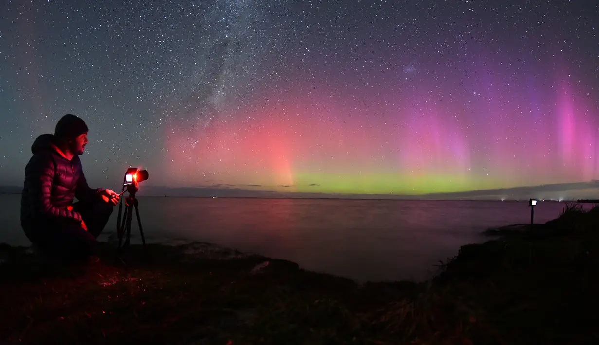 Seorang pria memotret Aurora Australis saat bersinar di cakrawala di atas perairan Danau Ellesmere di pinggiran Christchurch, Selandia Baru pada 24 April 2023. (AFP/Sanka Vidanagama)