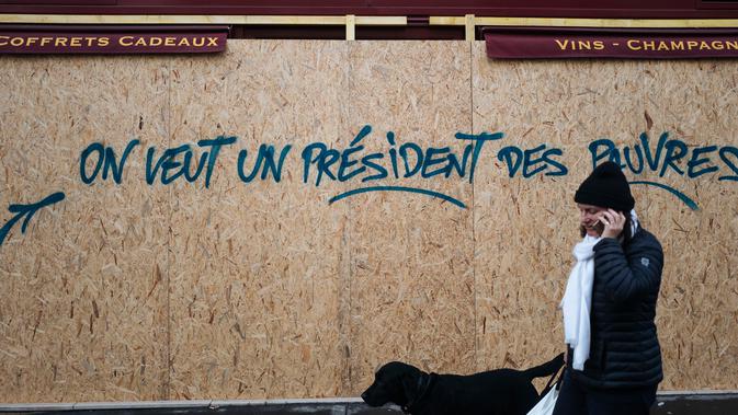 Seorang wanita berjalan melewati grafiti bertuliskan 'kami ingin seorang presiden untuk orang miskin' di sebuah toko dekat Arc de Triomphe, Paris, Prancis,  Minggu (2/12). Tiga orang tewas dalam unjuk rasa sejak dua pekan lalu. (AP Photo/Kamil Zihnioglu)