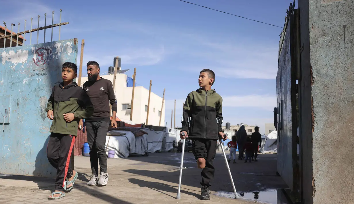Saat ini, Rateb Mahmoud Qaliq berusia sembilan tahun. Tampak dalam foto, Rateb Mahmoud Qaliq (kanan) berjalan menggunakan kruk di depan gerbang sekolah UNRWA yang telah menjadi tempat penampungan bagi pengungsi Palestina di Deir al-Balah, di Jalur Gaza tengah, pada 3 Desember 2025. (Bashar Taleb/AFP)