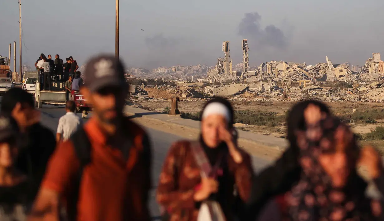 Asap membubung di atas bangunan-bangunan yang hancur saat warga Palestina meninggalkan kota Gaza ke arah selatan dengan membawa barang-barang mereka, melalui jalan pesisir dekat kamp pengungsi Nuseirat di bagian tengah Jalur Gaza, pada 12 September 2025. (Eyad BABA/AFP)