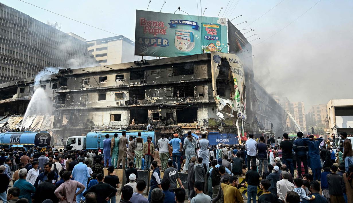Hingga Senin (19/1/2026), upaya pendinginan bangunan yang terbakar masih berlangsung. Tampak dalam foto, warga menyaksikan petugas pemadam kebakaran melakukan operasi pendinginan pada bangunan yang terbakar setelah kebakaran besar di sebuah pusat perbelanjaan di Karachi pada Senin 19 Januari 2026. (Asif HASSAN/AFP)