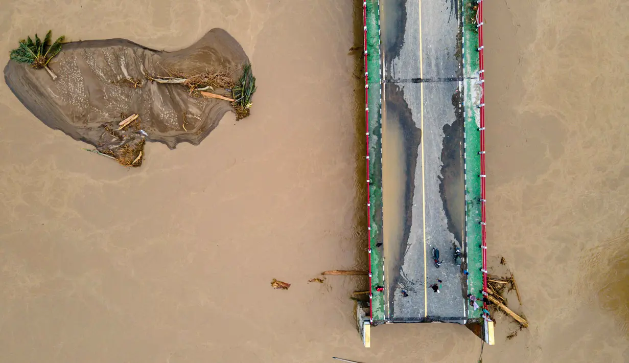 Kondisi sungai dipenuhi arus deras dengan air berwarna kuning pekat dan bergulung-gulung. Tampak foto udara menunjukkan jembatan yang rusak akibat banjir bandang di jalan utama yang menghubungkan Aceh dan Sumatera Utara di Meureudu, Kabupaten Pidie Jaya, Provinsi Aceh, Indonesia, pada Jumat 28 November 2025. (CHAIDEER MAHYUDDIN/AFP)