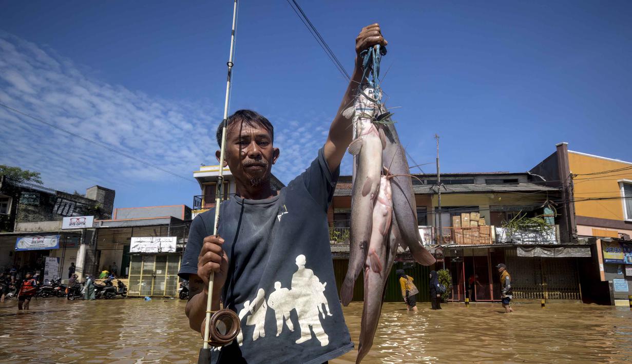 Air merendam permukiman hingga jalan raya dengan ketinggian beragam. Tampak dalam foto, seorang pria menunjukkan ikan tangkapannya saat memancing di jalanan Bandung yang tergenang banjir, Jawa Barat, pada Jumat 5 Desember 2025. (TIMUR MATAHARI/AFP)