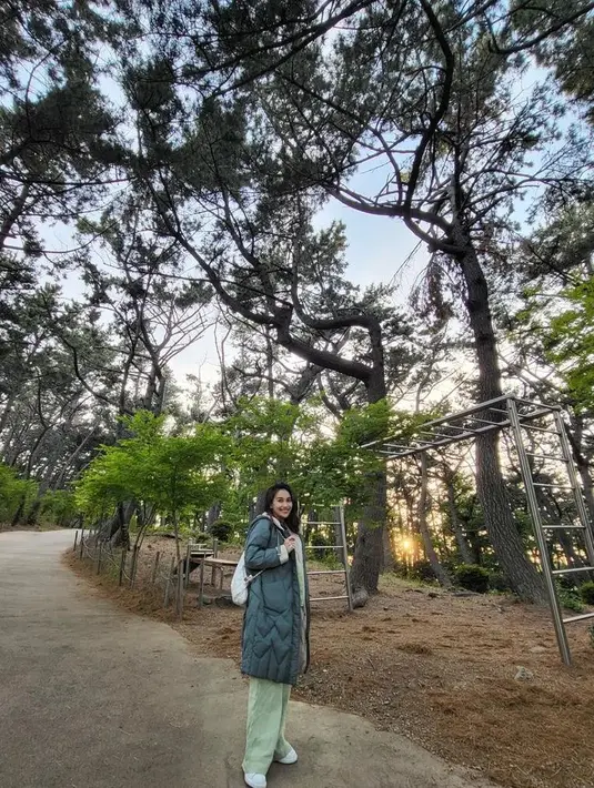Ia memakai set celana dan kemeja lengan panjang warna hijau mint dipadukan dengan mantel warna hijau tua. Terlihat menyatu dengan latar pepohonan di belakangnya.  [Foto: IG/ayutingting92].