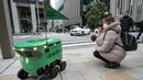 <p>Seorang wanita yang menggendong bayinya mengambil foto saat robot tanpa awak melewati mereka di sepanjang jalan saat uji coba layanan pengiriman robot oleh Uber Eats Jepang, di pusat kota Tokyo pada tanggal 5 Maret 2024. (Richard A. Brooks/AFP)</p>