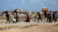 Otoritas Gaza menyeru kepada dunia internasional untuk menekan Israel agar menghentikan agresi serta membuka jalur bantuan tanpa hambatan. Tampak dalam foto, warga Palestina, membawa tas dan kardus kosong, berjalan melewati bangunan yang hancur saat mereka kembali dari titik distribusi makanan yang dikelola oleh Yayasan Kemanusiaan Gaza (GHF) yang didukung AS dan Israel, di dekat koridor Netsarim di Jalur Gaza tengah pada 5 Oktober 2025. (Eyad BABA/AFP)
