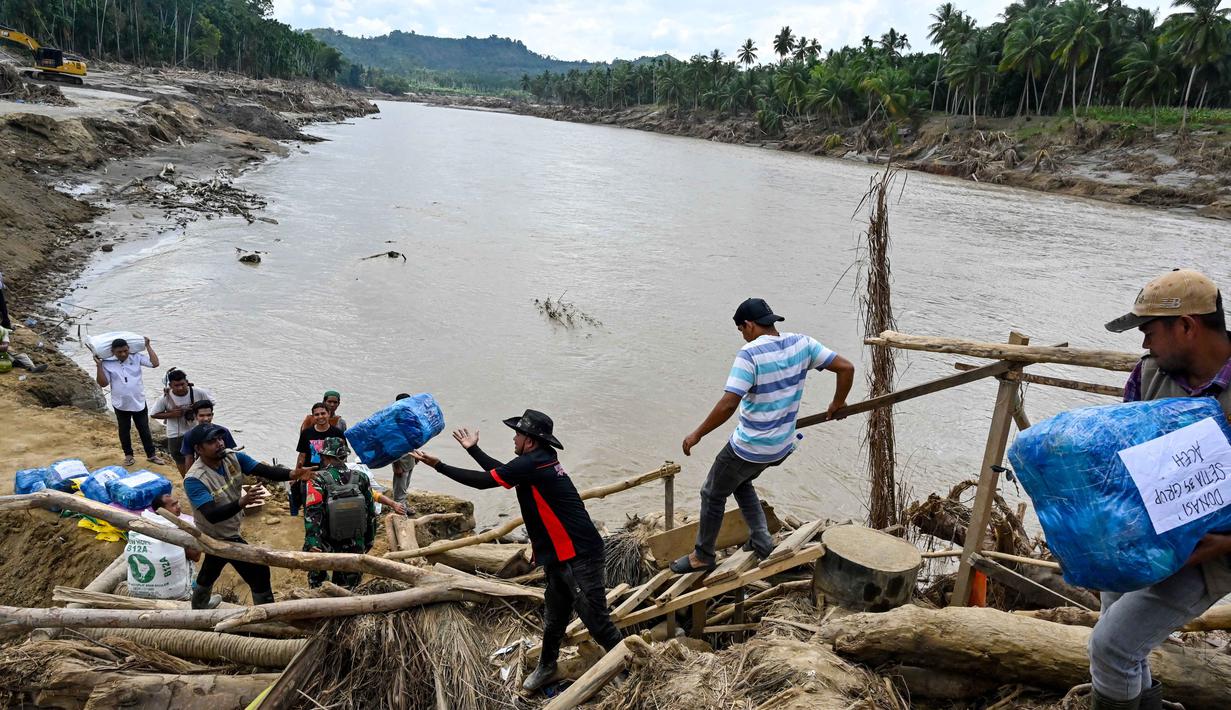 Salah satunya dengan melintasi tumpukan puing kayu untuk naik ke bagian jembatan yang masih utuh dan kokoh untuk dilewati. Tampak dalam foto, warga memindahkan bantuan logistik dengan melintasi sungai di jembatan yang baru dibangun yang menghubungkan Aceh dan Provinsi Sumatera Utara setelah jembatan tersebut hancur akibat banjir bandang di Sungai Peusangan, Bireuen, Provinsi Aceh, Selasa 9 Desember 2025. (CHAIDEER MAHYUDDIN/AFP)