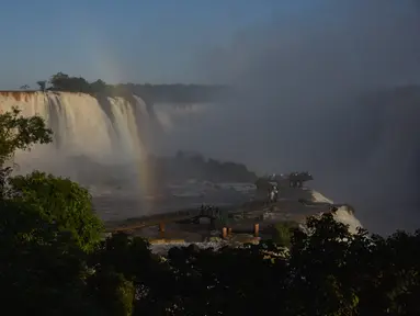 Pengunjung berjalan di sepanjang jalan setapak saat pelangi muncul di depan sebagian pemandangan Air Terjun Devil's Throat, bagian dari sistem Air Terjun Iguazu di Sungai Iguazu di Taman Nasional Iguacu, Negara Bagian Parana, dekat Foz do Iguacu di Brasil selatan yang berbatasan dengan Argentina, saat matahari terbenam pada 21 Mei 2024. (Mariana SUAREZ / AFP)
