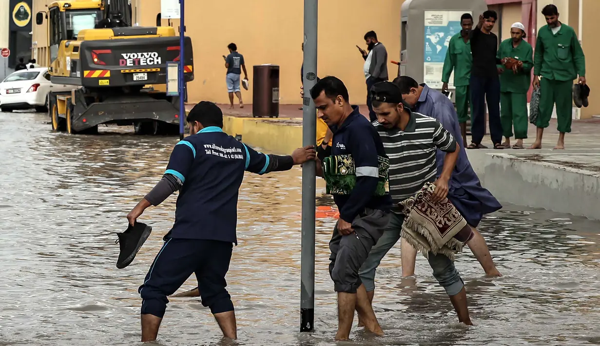 Pemerintah Dubai, UEA, mendesak warga untuk tetap berada di dalam rumah, kecuali jika benar-benar diperlukan. Tampak dalam foto, para pria mengangkat ujung celana mereka untuk menyeberangi genangan air banjir saat melintasi jalan setelah hujan lebat, di Dubai, Uni Emirat Arab (UEA) pada Jumat 19 Desember 2025. (FADEL SENNA/AFP)