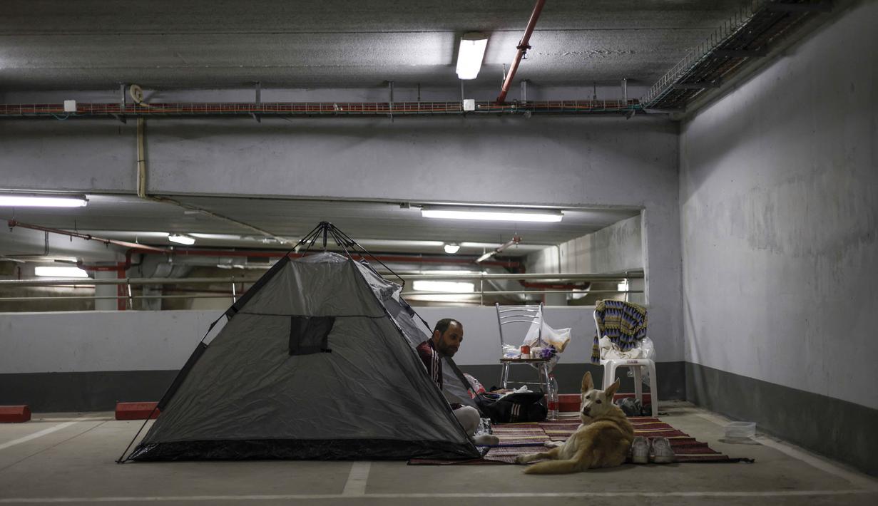 Wilayah udara Israel juga sempat ditutup total untuk penerbangan sipil guna mengantisipasi ancaman rudal balistik dan drone dari Teheran. Tampak dalam foto, seorang pria bersama anjingnya berlindung di tempat parkir bawah tanah di Haifa pada Minggu 1 Maret 2026, di tengah kekhawatiran akan serangan Iran. (Jalaa MAREY/AFP)