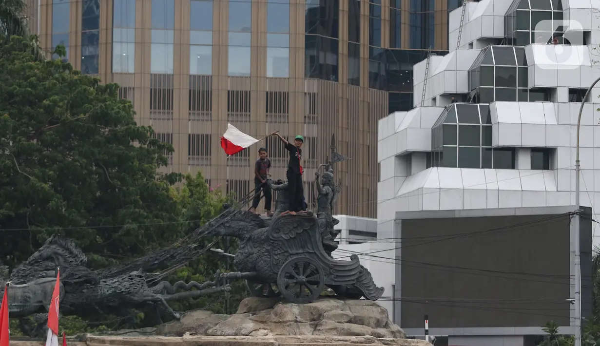Massa aksi demo tolak omnibus law UU Cipta Kerja naik ke atas patung arjuna wiwaha mengibarkan bendera merah putih di Jalan Medan Merdeka Barat, Jakarta Pusat, Selasa (20/10/2020). Aksi mereka lantas disambut meriah oleh para massa yang masih bertahan. (Liputan6.com/Herman Zakharia)