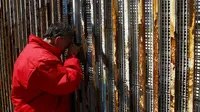 Seorang warga Meksiko terbujur kaku sambil memegang pagar perbatasan sambil berbincang dengan keluarganya di Playas Tijuana, Meksiko, (12/4). Tembok ini memisahkan antara Meksiko dan Amerika Serikat. (REUTERS / Jorge Duenes)