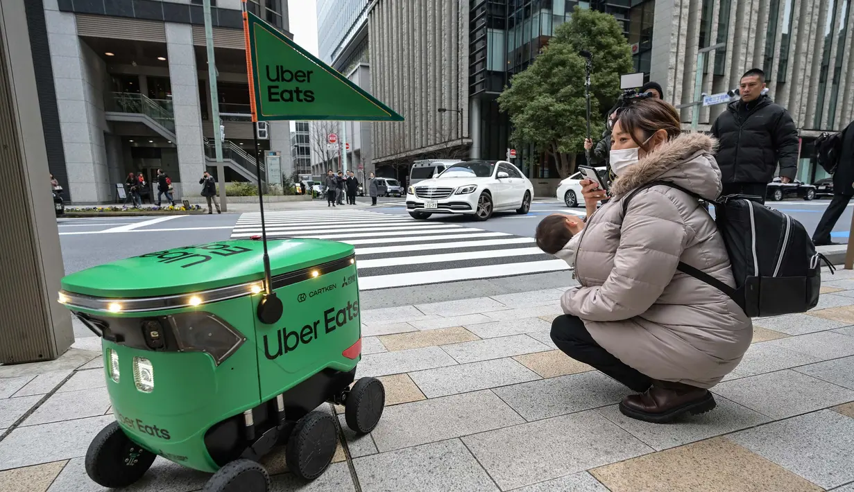 <p>Seorang wanita yang menggendong bayinya mengambil foto saat robot tanpa awak melewati mereka di sepanjang jalan saat uji coba layanan pengiriman robot oleh Uber Eats Jepang, di pusat kota Tokyo pada tanggal 5 Maret 2024. (Richard A. Brooks/AFP)</p>