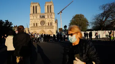 Seorang perempuan melintasi orang-orang yang berhenti di depan Katedral Notre Dame saat kembali membunyikan lonceng utamanya di Paris, Rabu (15/4/2020). Lonceng utama katedral kembali dibunyikan setelah satu tahun mengalami kebakaran yang sampai mengejutkan seluruh warga dunia. (AP/Christophe Ena)