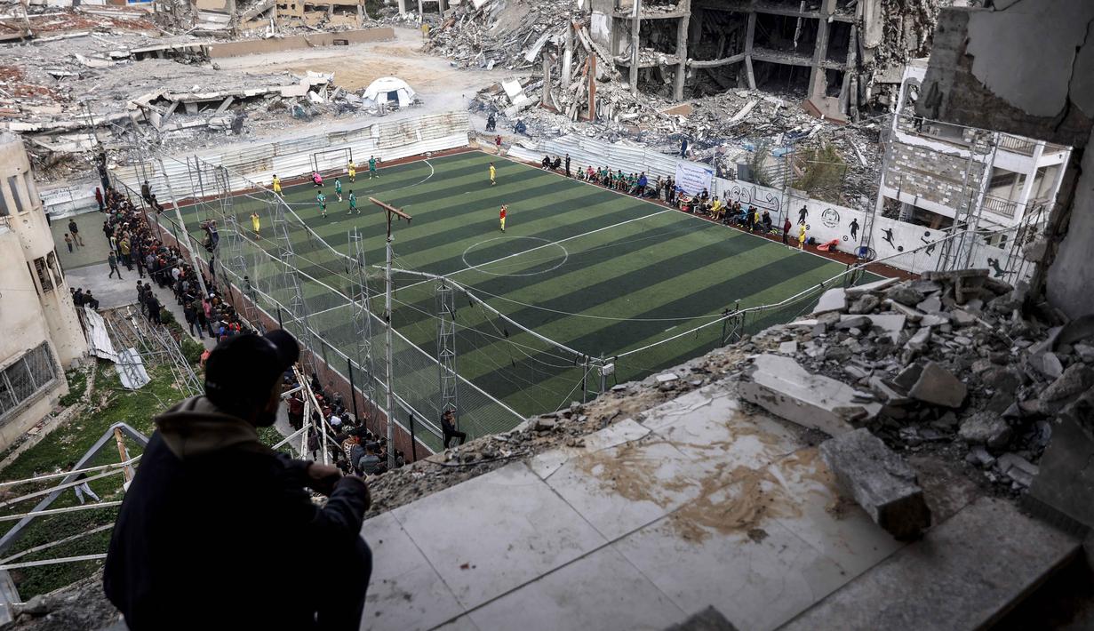 Warga Palestina menyaksikan pertandingan sepak bola lima lawan lima di tengah reruntuhan bangunan yang hancur di Kota Gaza pada Senin 9 Februari 2026. Keriuhan memecah kesunyian di antara reruntuhan bangunan di Kota Gaza. (Omar AL-QATTAA/AFP)