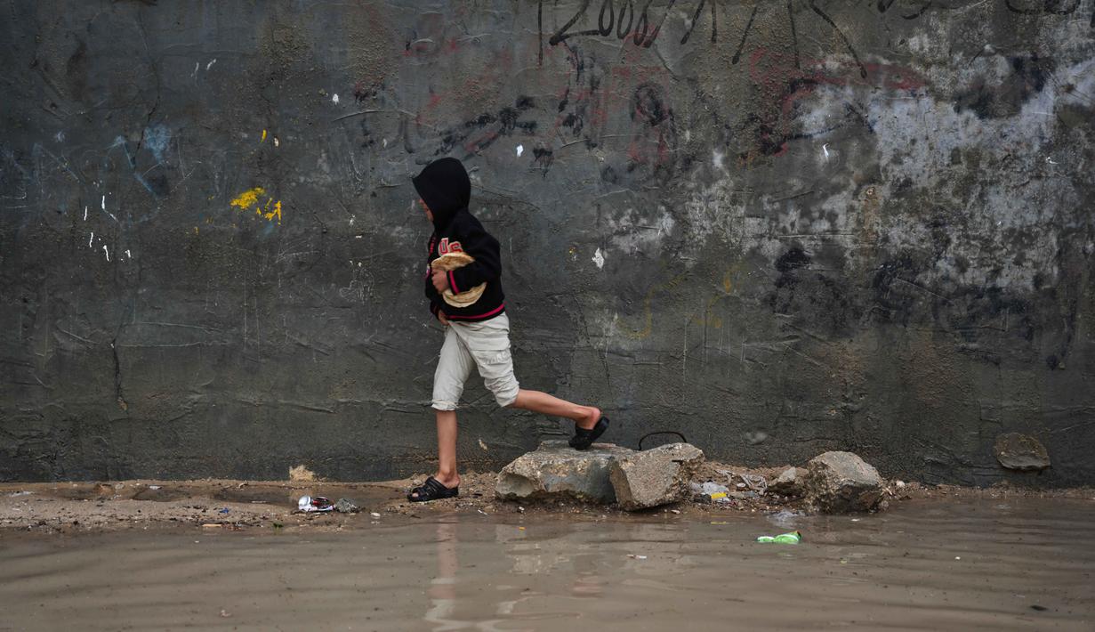 Situasi ini meningkatkan risiko penyebaran penyakit menular, terutama bagi anak-anak dan lanjut usia (lansia). Tampak dalam foto, seorang anak pengungsi Palestina berjalan melewati kamp tenda yang basah kuyup setelah hujan lebat di Deir al-Balah, Jalur Gaza tengah, Kamis, 26 Maret 2026. (AP Photo/Abdel Kareem Hana)