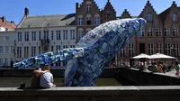 Wisatawan duduk dekat patung paus dari 5 ton sampah plastik yang ditampilkan di sungai Brugges, Belgia, Sabtu (14/7). Patung paus tersebut bertujuan untuk menyoroti ancaman yang diakibatkan oleh penggunaan plastik secara besar-besaran. (AFP/JOHN THYS)