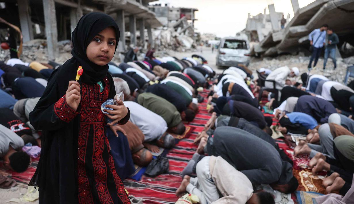 Seorang gadis memegang sepotong permen berdiri di antara warga Palestina yang melaksanakan salat menandai dimulainya Idul Adha, Hari Raya Kurban, di jalan yang rusak parah di Khan Yunis, Jalur Gaza Selatan, pada Rabu 27 Mei 2026. (BASHAR TALEB/AFP)