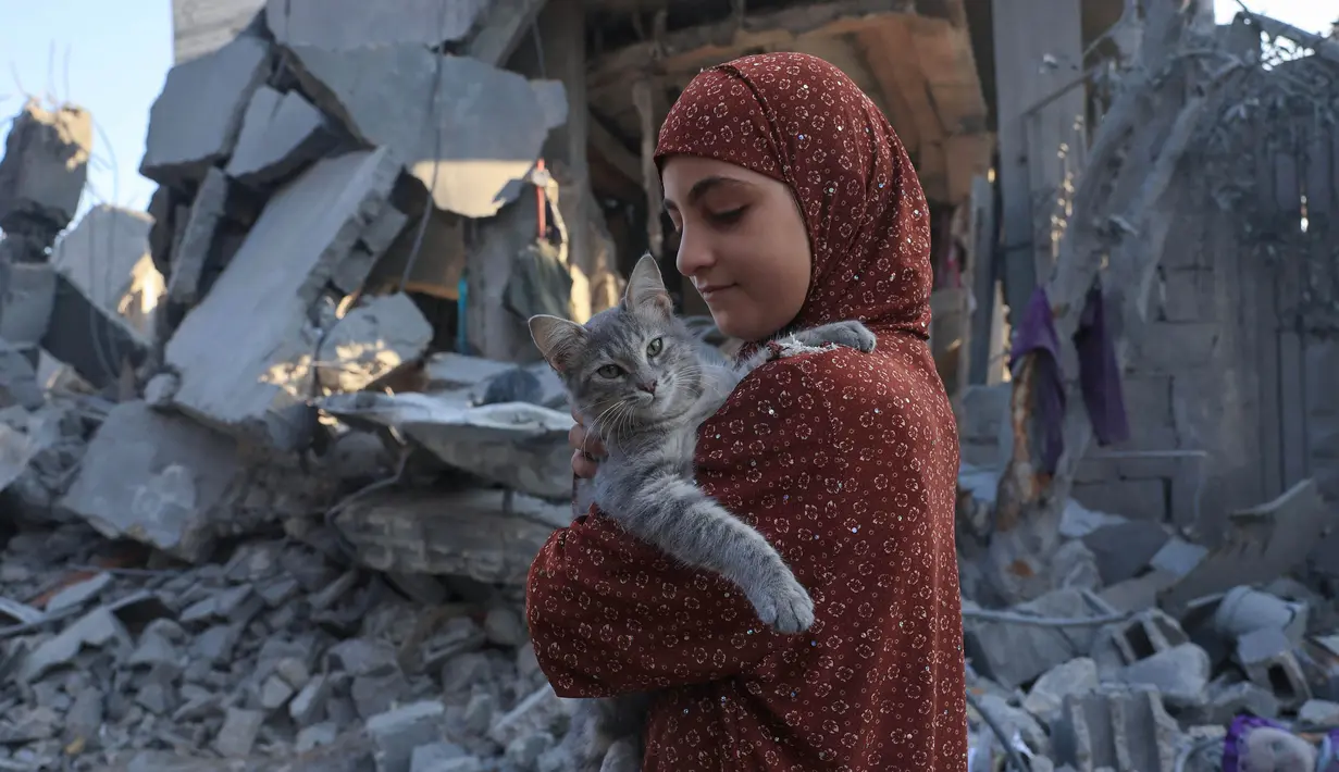 Seorang anak perempuan Palestina menggendong seekor kucing saat dia memeriksa kerusakan setelah pemboman Israel di Rafah di Jalur Gaza selatan, pada 18 Oktober 2023. (SAID KHATIB/AFP)