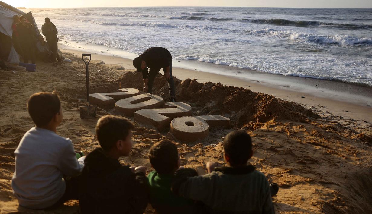Cuaca ekstrem yang ditandai dengan hujan deras, angin kencang, dan suhu dingin memperburuk kondisi pengungsi Palestina di Gaza hingga menimbulkan angka kematian baru. Tampak dalam foto, anak-anak menyaksikan seniman Yazed Abo Jarad membuat patung pasir untuk tahun mendatang di Deir El-Balah, Jalur Gaza tengah pada 30 Desember 2025. (Bashar Taleb/AFP)