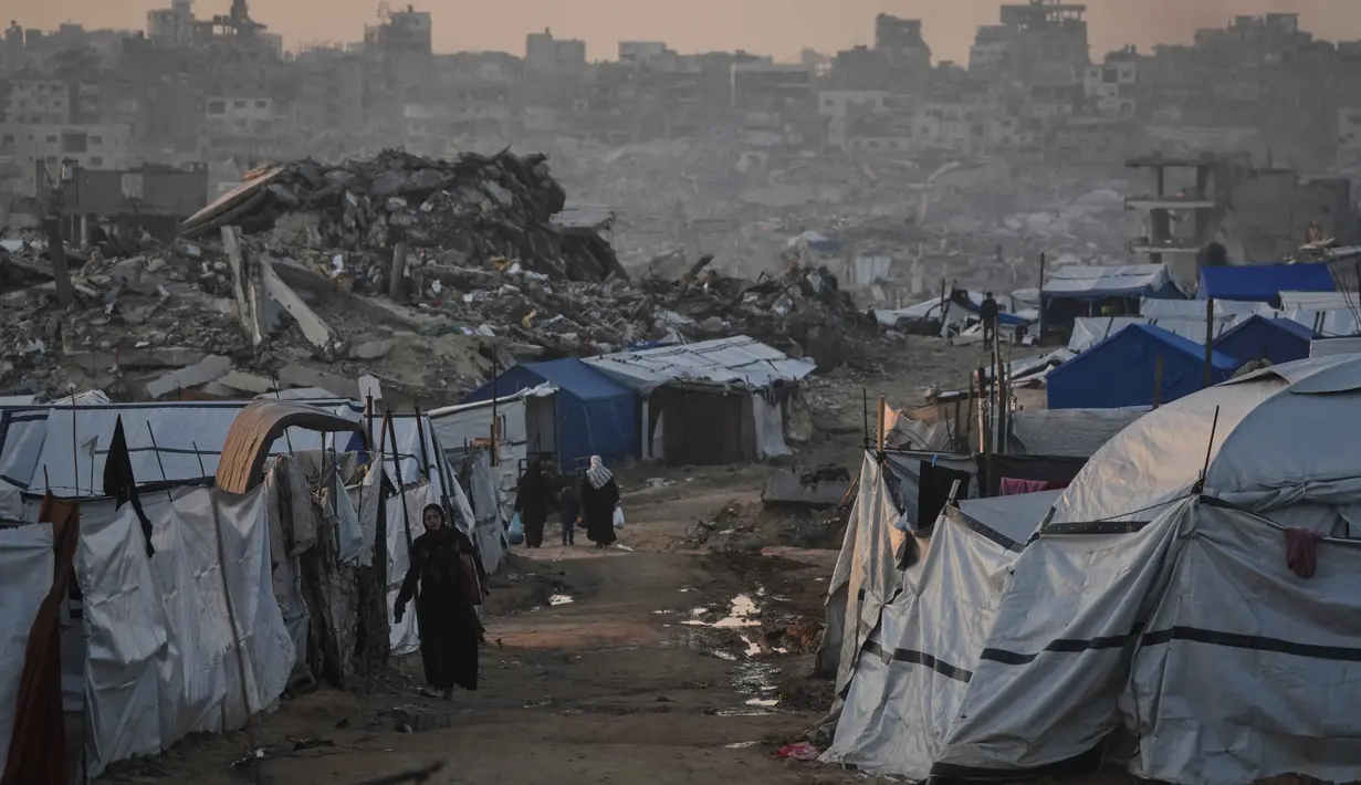 Saat ini, ratusan palet bantuan berisi peralatan medis dan kebutuhan pokok telah disiapkan di Yordania dan Mesir. Tampak dalam foto, warga Palestina berjalan melewati perkemahan tenda di tengah bangunan-bangunan yang hancur akibat serangan udara dan darat Israel di Kota Gaza, Selasa 6 Januari 2026. (AP Photo/Jehad Alshrafi)